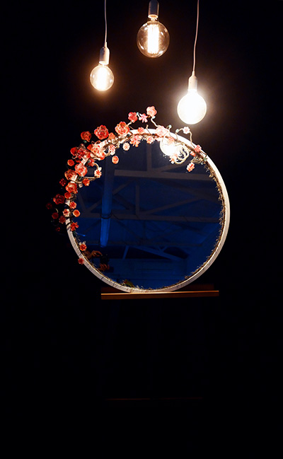 An installation featuring a round mirror with small flowers on its rim, sitting on an easel illuminated by 3 hanging lights in a dimmed environment.