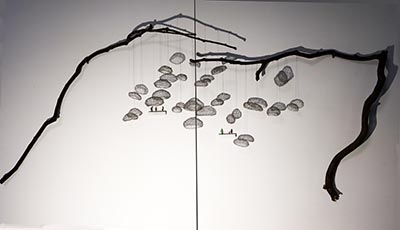 A sculpture features wire shapes dangling from two separate tree branches, accompanied by miniature human figures, all mounted on a wall.