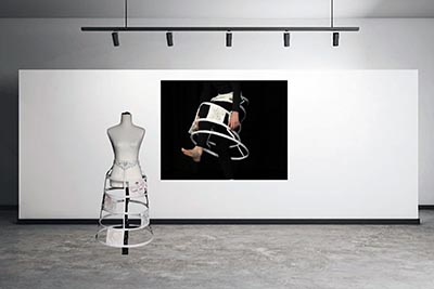 Installation featuring a mannequin torso with a bell-shaped cage crinoline in front of a close-up image of someone attempting to fit into one.