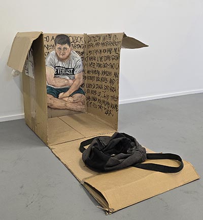 The painting depicts a person seated, set against a backdrop consisting of text that fills the inner surfaces of a cardboard box. A black bag is observed at the forefront, resting on a cardboard box.