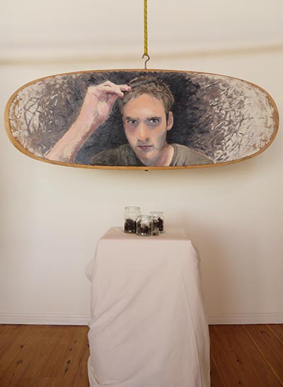 An installation features an oval-shaped portrait painting of a person combing their hair, framed by a light brown border. In front of it, there are a few jars displayed on a plinth.