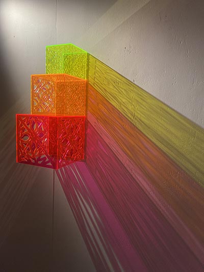 A sculpture consists of 3 cubes arranged from smallest to largest, sitting on top of each other. The top cube is yellowish-green, followed by an orange cube, and then a red cube at the base.