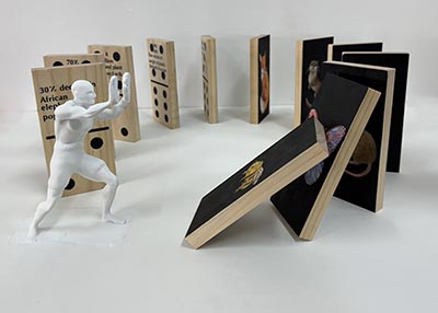 An installation featuring a human figurine in a pushing stance, with the first two dominos tipping over. The dominos, arranged in a circular formation, are painted and include text on their surfaces.