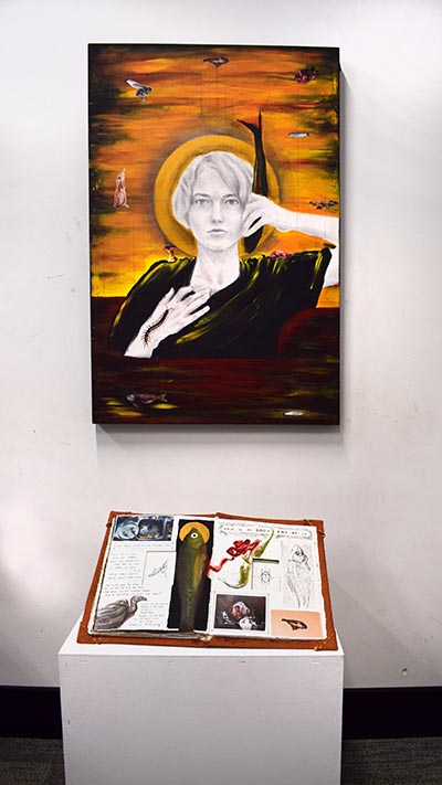 An installation comprising a painting displayed on the wall, with a book of collages placed on a plinth directly beneath it.