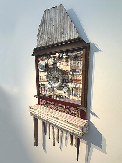 Sculpture displayed on the wall, made from materials including wood, chair/table legs, corrugated iron, a rug, kitchen utensils, and other elements
