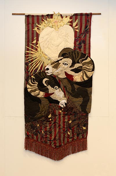 A wall-hanging textile artwork featuring a golden Sacred Heart,  rams and other elements.