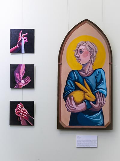 On the right, a self-portrait painting of the artist holding a golden rabbit is displayed in a cathedral-style frame. On the left, three vertically arranged artworks depict images related to hands engaged in spiritual practices.