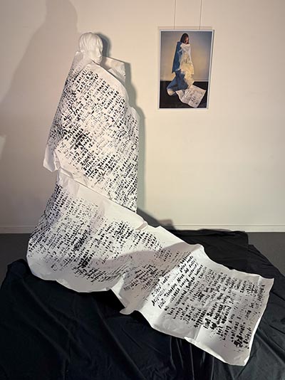 An installation showcasing a life-size human sculpture wrapped in paper adorned with printed words, displayed alongside an image of a real person similarly wrapped in the same paper.