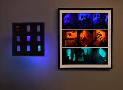 A sculpture featuring a square structure with nine cut-out openings arranged in a 3x3 grid, illuminated from behind, placed beside a single frame containing three  photographs.