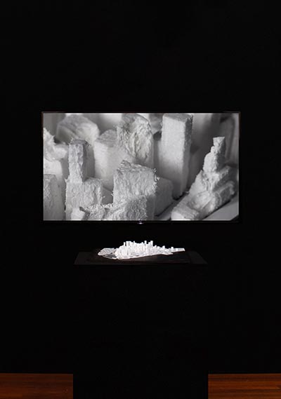 An installation showcasing a  sculpture of a city carved from styrofoam and displayed on a plinth, positioned in front of a close-up photograph highlighting a specific section of the sculpture.