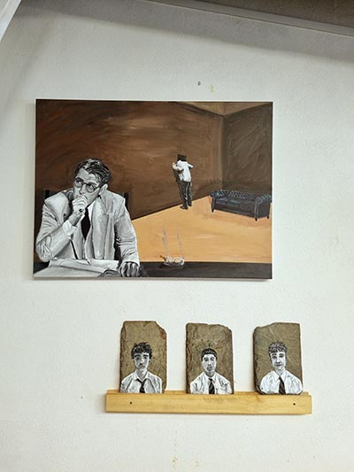 A large painting hangs on the wall, with 3 small individual portraits of young males leaning against the wall on a wall-mounted shelf directly beneath it.