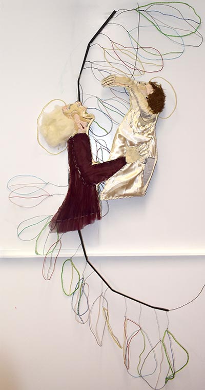 A wall-mounted textile sculpture depicting a human figure holding and lifting another human figure in the air. The background features a long, black, curving stem with multiple leaf-shaped outlines extending outward.