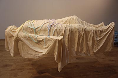 A sculpture depicting a human figure in a lying position, covered in beige, with coloured accents highlighting some of the creases on the upper torso. The figure appears suspended in the air.