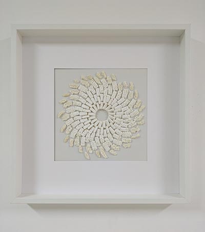 A sculpture of white rectangular chewing gums arranged in circular layers, with the inner layer intact and increasing deformation and cracks outward, culminating in complete distortion at the outermost layer. The artwork is framed and displayed on a wall.