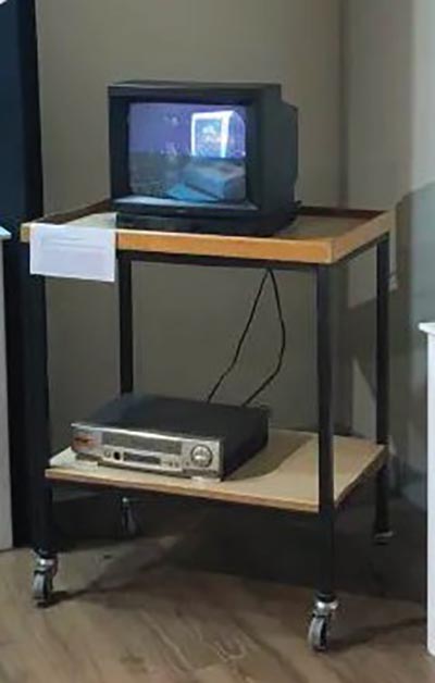 An installation featuring a cathode ray tube (CRT) television, displaying visuals, placed on the top shelf of a wheeled two-shelf unit, with a videocassette recorder (VCR) connected and positioned on the bottom shelf.