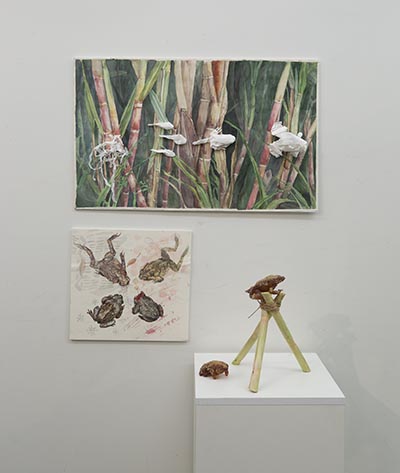 An installation featuring a large painting of sugarcanes with toad sculptures on top, a smaller painting of four toads, and a sculpture of a toad atop tied sugarcanes forming a triangular pod, with another toad nearby. The sculpture sits on a plinth in front of the wall-mounted paintings.