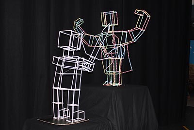 An installation featuring two geometric wireframe human-like sculptures against a black background: one, in white, holding a sword at a lower level; the other, in coloured outlines, wearing a cape at a higher level and flexing its muscles to display strength.