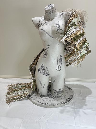 A mannequin torso featuring imageries and text around the neck sits atop a round base with imagery and text that share a cohesive visual connection with the torso. A woven textile drapes from the left shoulder, wrapping around the back and extending to the right side of the torso.