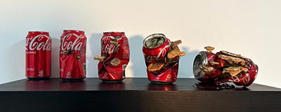 An installation of 5 Coca-Cola cans displayed in varying conditions: the first can is untouched, while the second shows dents and fungi growth. Progressively, the cans exhibit more prominent dents, increased fungi presence and larger fungal growths.