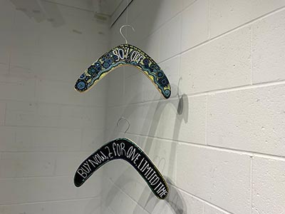 Two boomerangs featuring Indigenous art, each with a clothes hanger hook attached, suspended vertically. The top boomerang has '50% OFF' written on it, while the bottom one displays 'BUY NOW, 2 FOR ONE, LIMITED TIME.'