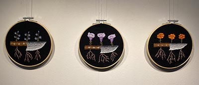 Three circular pieces of fabric feature embroidered designs of flowering plants growing on knives, with their roots extending below. Each circle showcases a different type of flower.