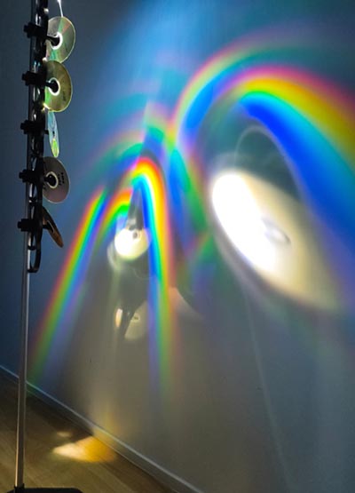 An installation featuring rainbow-coloured light reflections and disc reflections, created by light deflecting off 5 discs vertically attached to a pole.