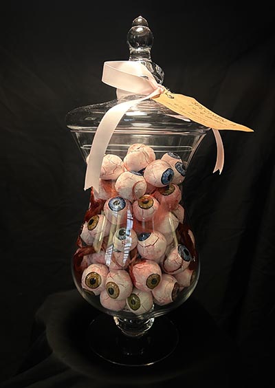 A sculpture depicting a glass container filled with eyeballs, adorned with a pink ribbon and a tag placed on top.